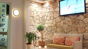 A reception area with a stone wall, a flat-screen TV, a sofa with patterned cushions, a glass coffee table with candles, and a potted plant. - Amaryllis Hotel Apartments (Nafplio)