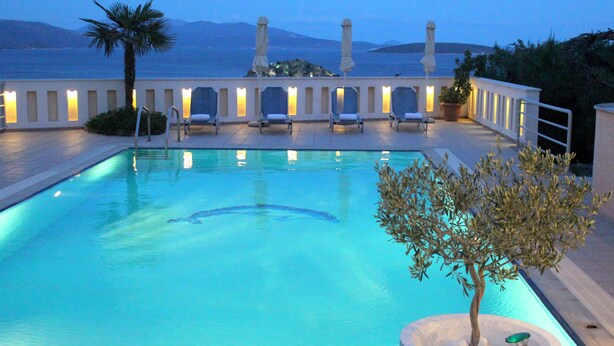 A pool area with lounge chairs and umbrellas, a palm tree, and a view of the sea and mountains. - Amaryllis Hotel Apartments (Nafplio)