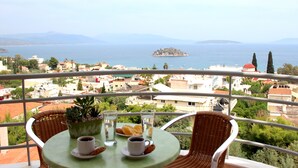 A balcony with a table set for two, overlooking a coastal town with buildings and a distant island. - Amaryllis Hotel Apartments (Nafplio)