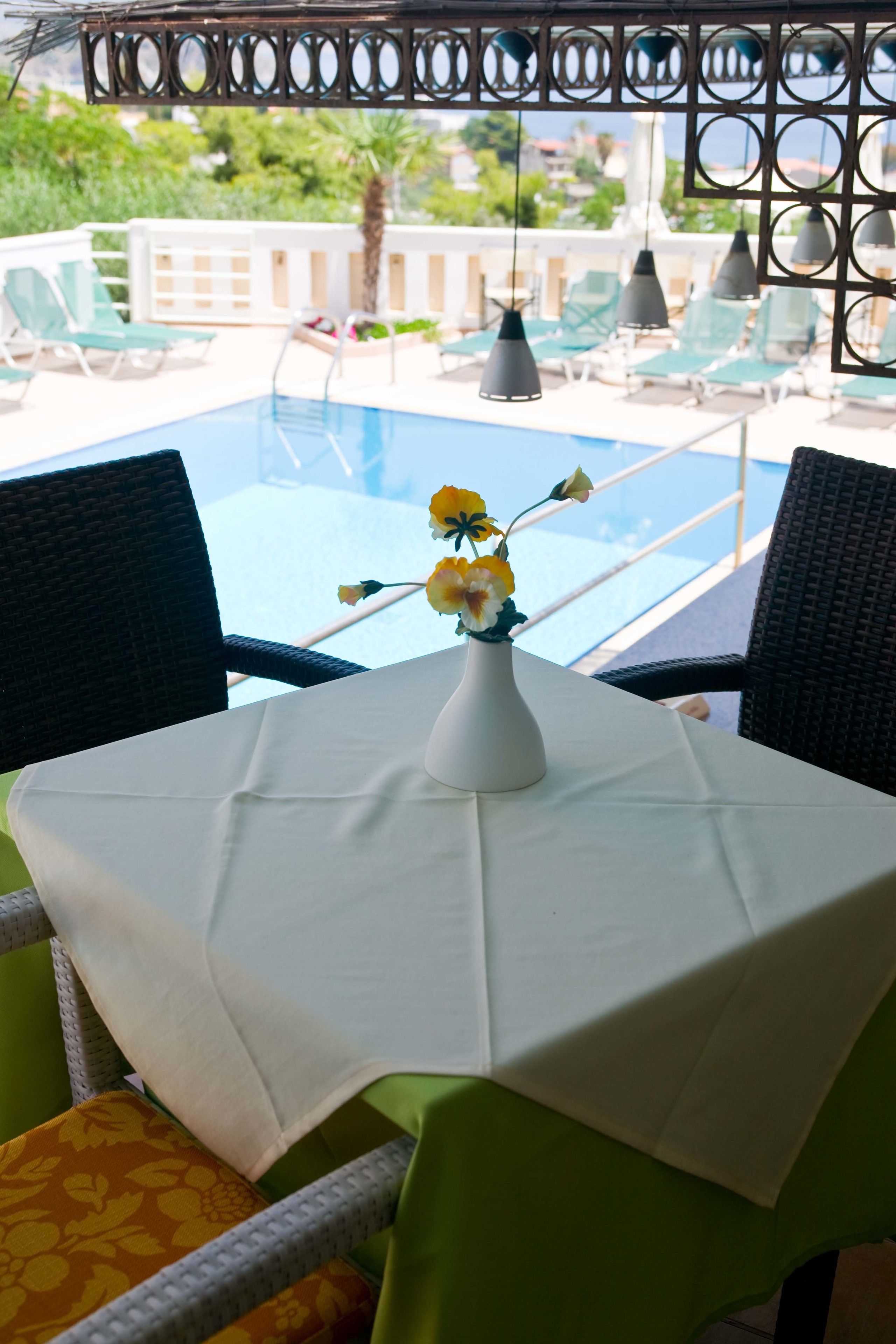 A table with a white tablecloth, a vase with yellow flowers, and a view of a pool and lounge chairs.