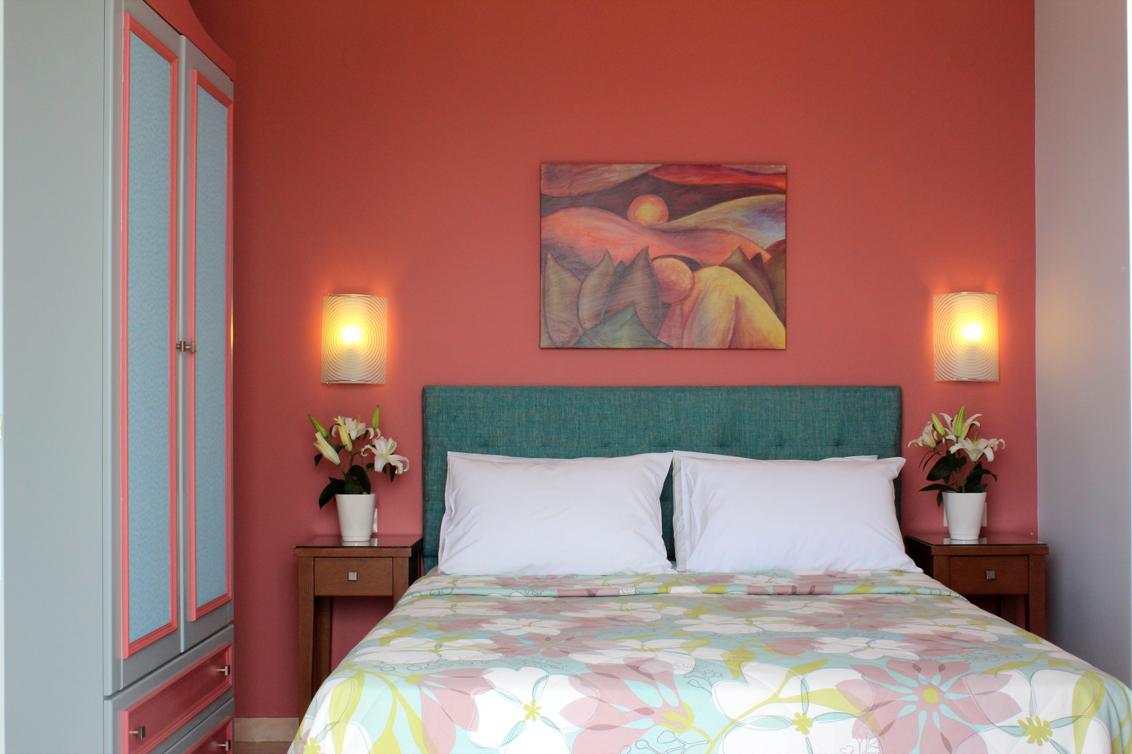 A bedroom with a teal headboard, two white pillows, a floral bedspread, a painting on the wall, and two bedside lamps.