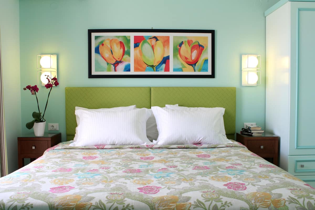 Ein Bett mit floralem Überwurf, zwei weiße Kissen und ein grünes Kopfteil. Darüber hängen drei gerahmte Blumenbilder.