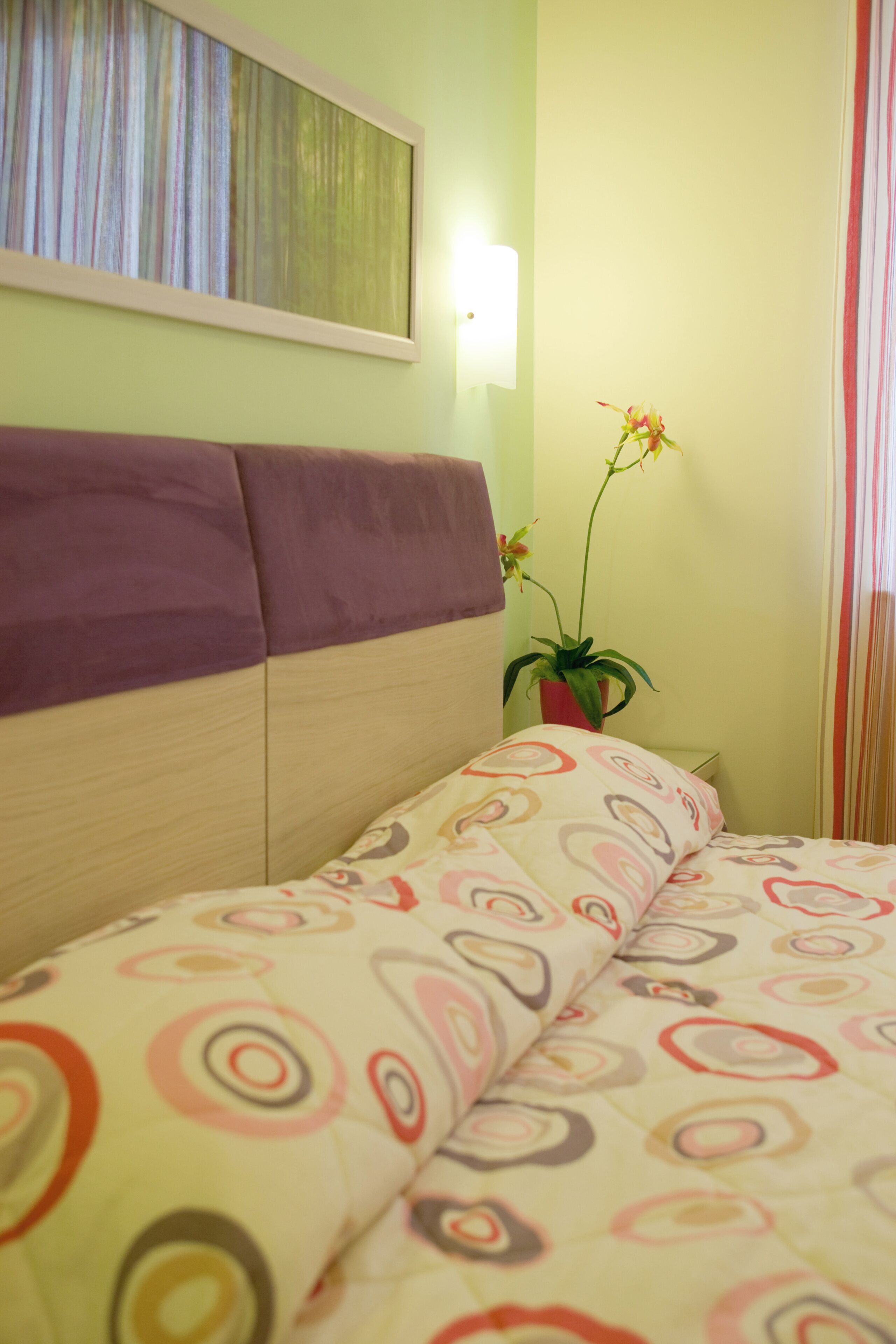 A bed with a patterned bedspread, a headboard, a wall-mounted light, and a potted plant.