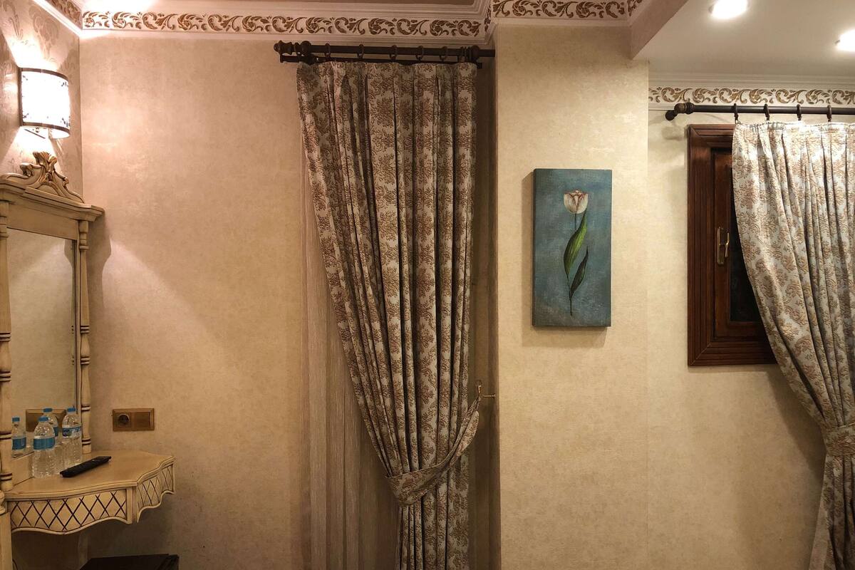 A hotel room with a bed, a bedside table with a lamp, a painting, and curtains.