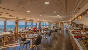 A restaurant with round tables and chairs, a buffet with various food items, and a view of the sea through large windows. - Kaftan Hotel (Istanbul)