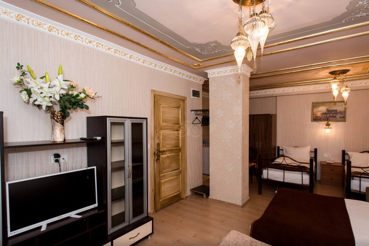 A hotel room with a television, a wooden door, a bed with two pillows, a nightstand, and a chandelier.