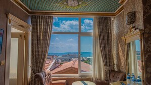 A room with a large window offering a view of rooftops and buildings, a small round table, two armchairs, and a mirror with a decorative frame. - Kaftan Hotel (Istanbul)