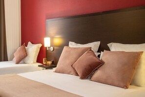 Habitación de hotel con dos camas, cada una con sábanas blancas y almohadas marrones, un cabecero de madera, una mesita de noche con lámpara y una pared de acento roja.