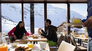 Une table dressée pour le petit-déjeuner avec vue sur les montagnes, des tables en bois et une variété d’aliments proposés.