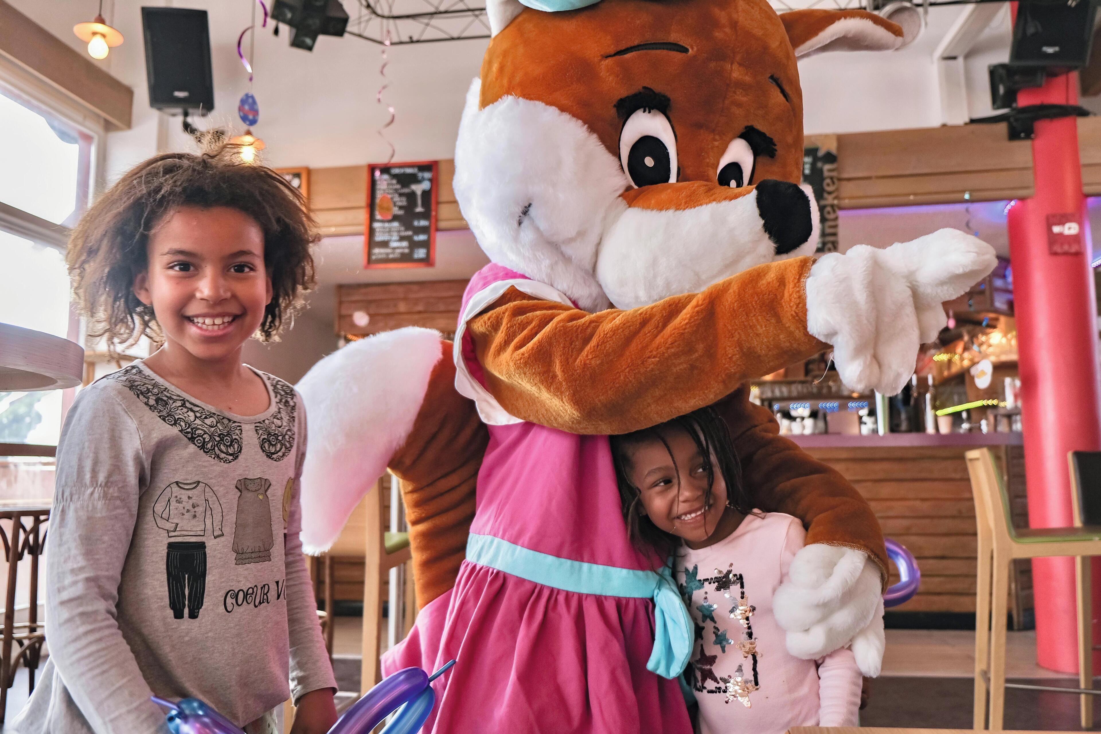 A person in a costume with a large character, a child, and a child in a pink dress in a room with wooden walls and a bar area in the background.