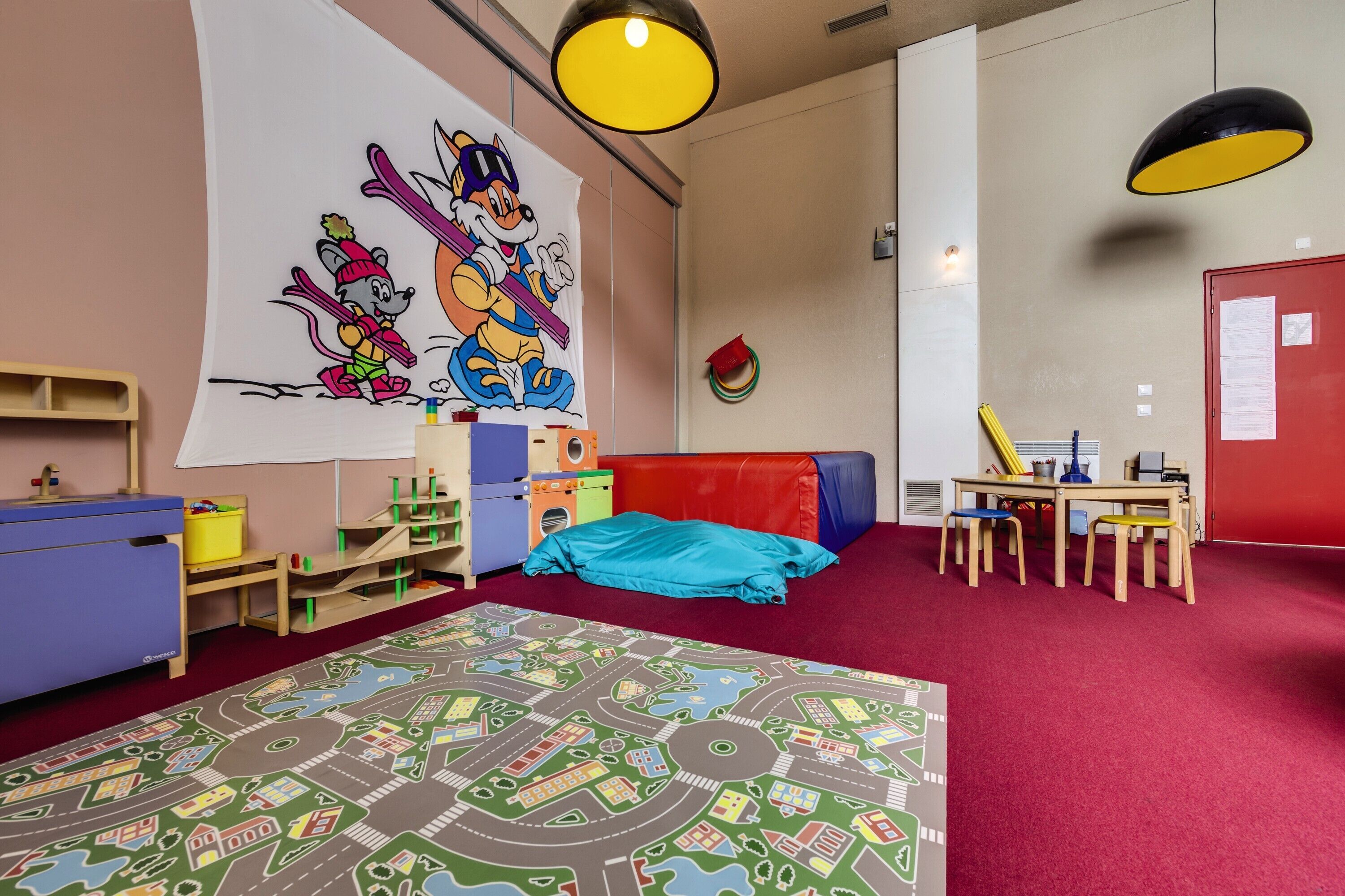 A children's playroom with a large cartoon mural, a red carpet, and a play area with a table and chairs.