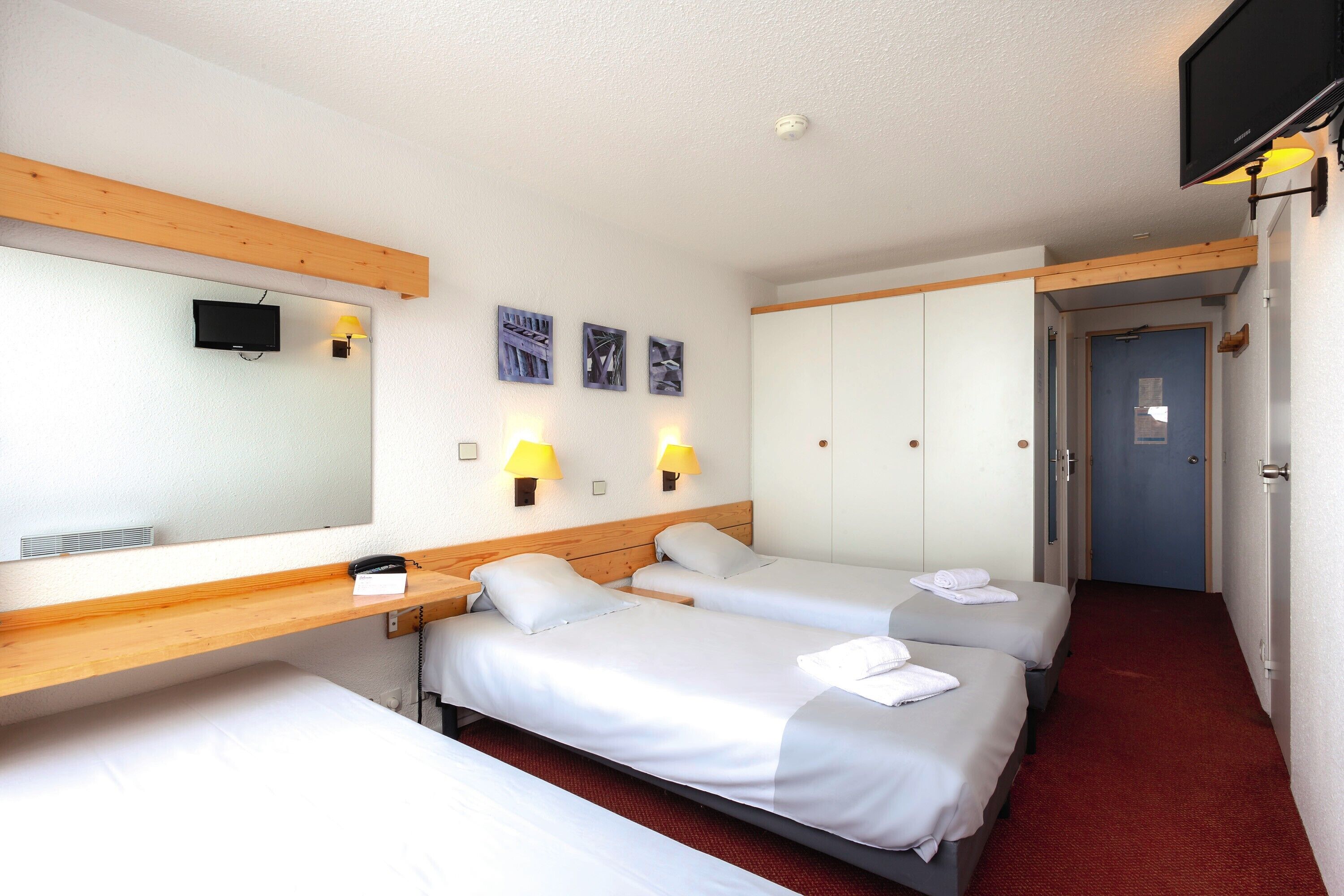 Une chambre d’hôtel avec deux lits, une tête de lit en bois, une télévision et des lampes fixées au mur.