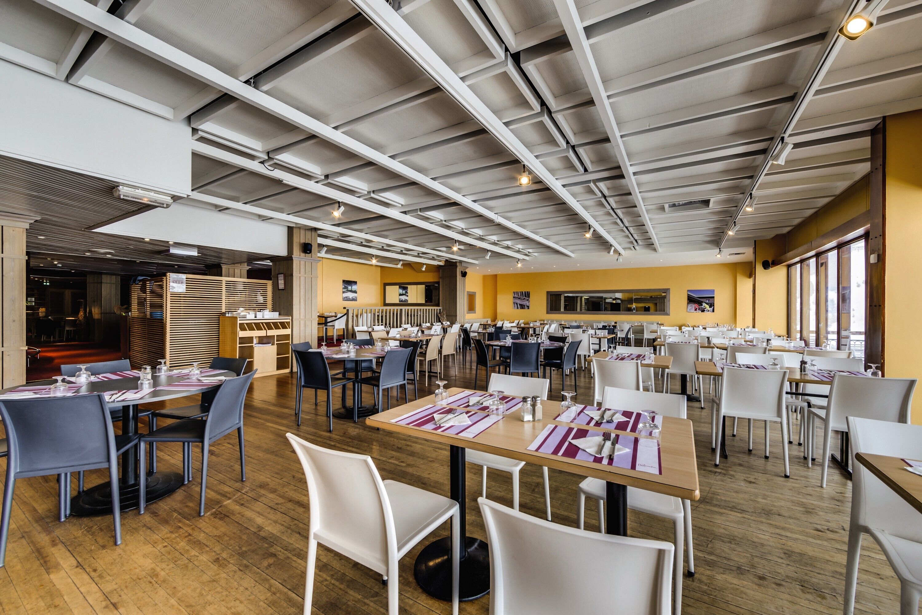 A spacious dining area with multiple tables and chairs, high ceilings with exposed beams, and large windows allowing natural light.
