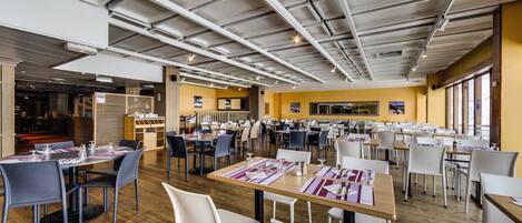 A spacious dining area with multiple tables and chairs, high ceilings with exposed beams, and large windows allowing natural light.