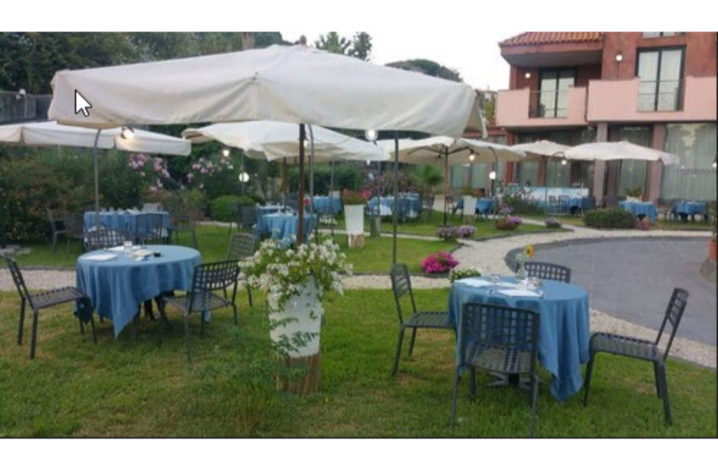 Area pranzo all'aperto con tavoli e sedie rotonde, coperta da ombrelloni bianchi, con sullo sfondo un edificio e vegetazione.