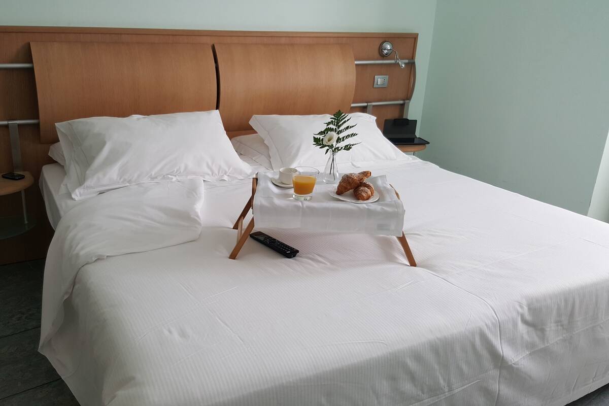 Ein ordentlich bezogenes Bett mit weißen Laken, ein Nachttisch mit Orangensaft, Croissants und einer Fernbedienung.