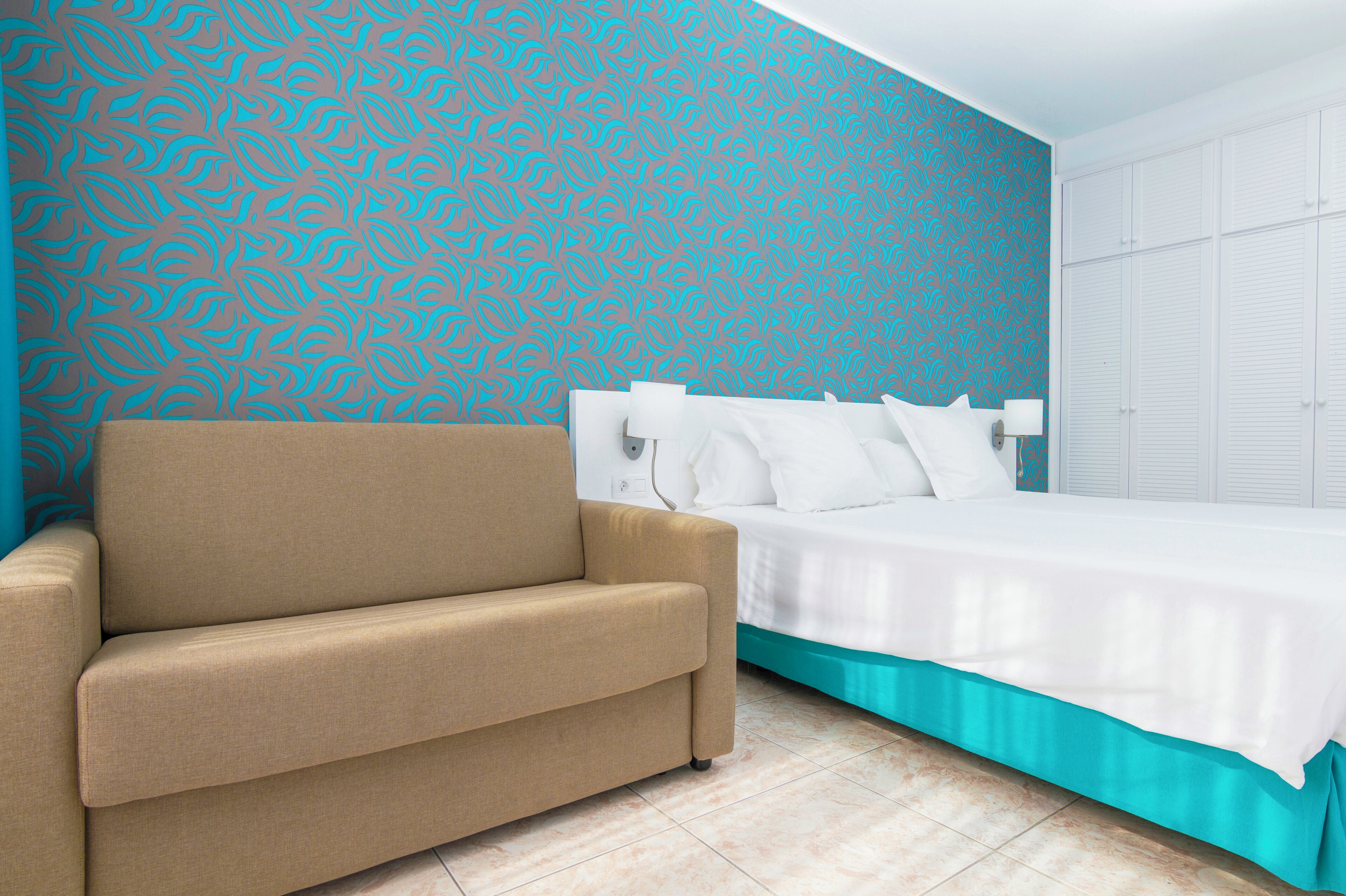 Un dormitorio con un sofá color beige, una cama con ropa de cama blanca y una pared con estampado azul.