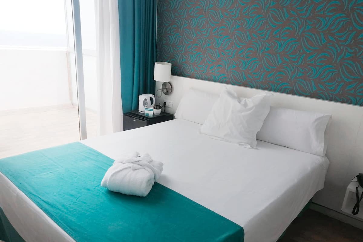Habitación de hotel con cama, mesita de noche, lámpara y vistas al exterior.