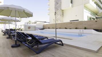 Un área junto a la piscina con sillones y sombrillas, piscina y un edificio moderno al fondo.