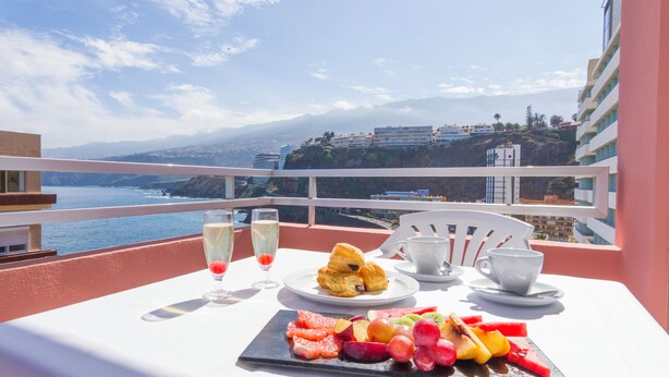 Een tafel met een fruitschaal, gebak en drankjes op een balkon met uitzicht op zee en gebouwen.