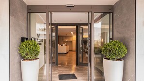 L’entrée d’un hôtel moderne, avec une porte en verre, un comptoir d’accueil et deux plantes en pot.