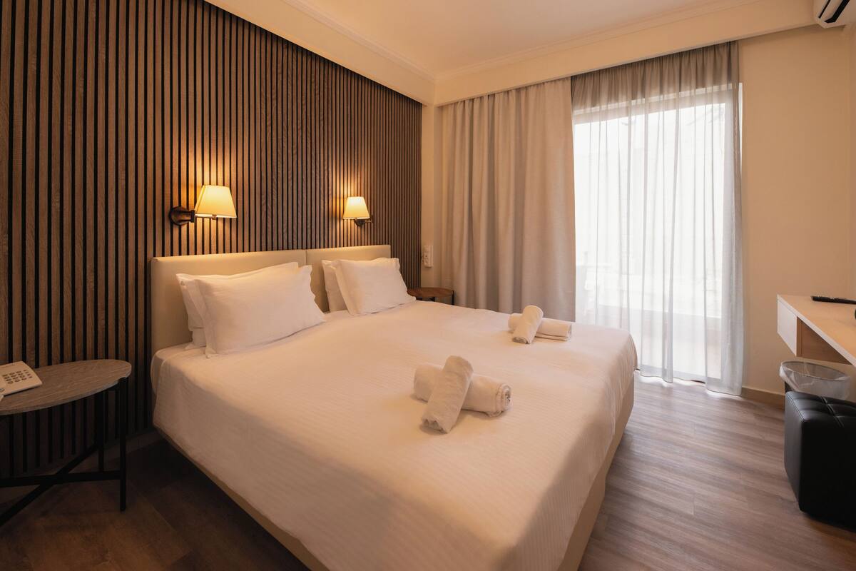 A hotel room with a large bed, two bedside lamps, a desk with a phone, and a window with curtains.
