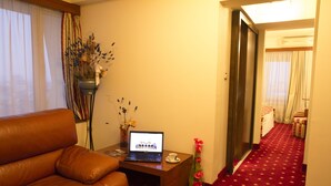 Une chambre d’hôtel avec un canapé en cuir marron, une table d’appoint en bois sur laquelle se trouve un ordinateur portable, un vase avec des fleurs et un couloir recouvert d’une moquette rouge.