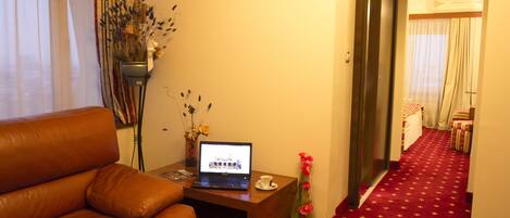 A hotel room with a brown leather sofa, a wooden side table with a laptop, a vase with flowers, and a red carpeted hallway.