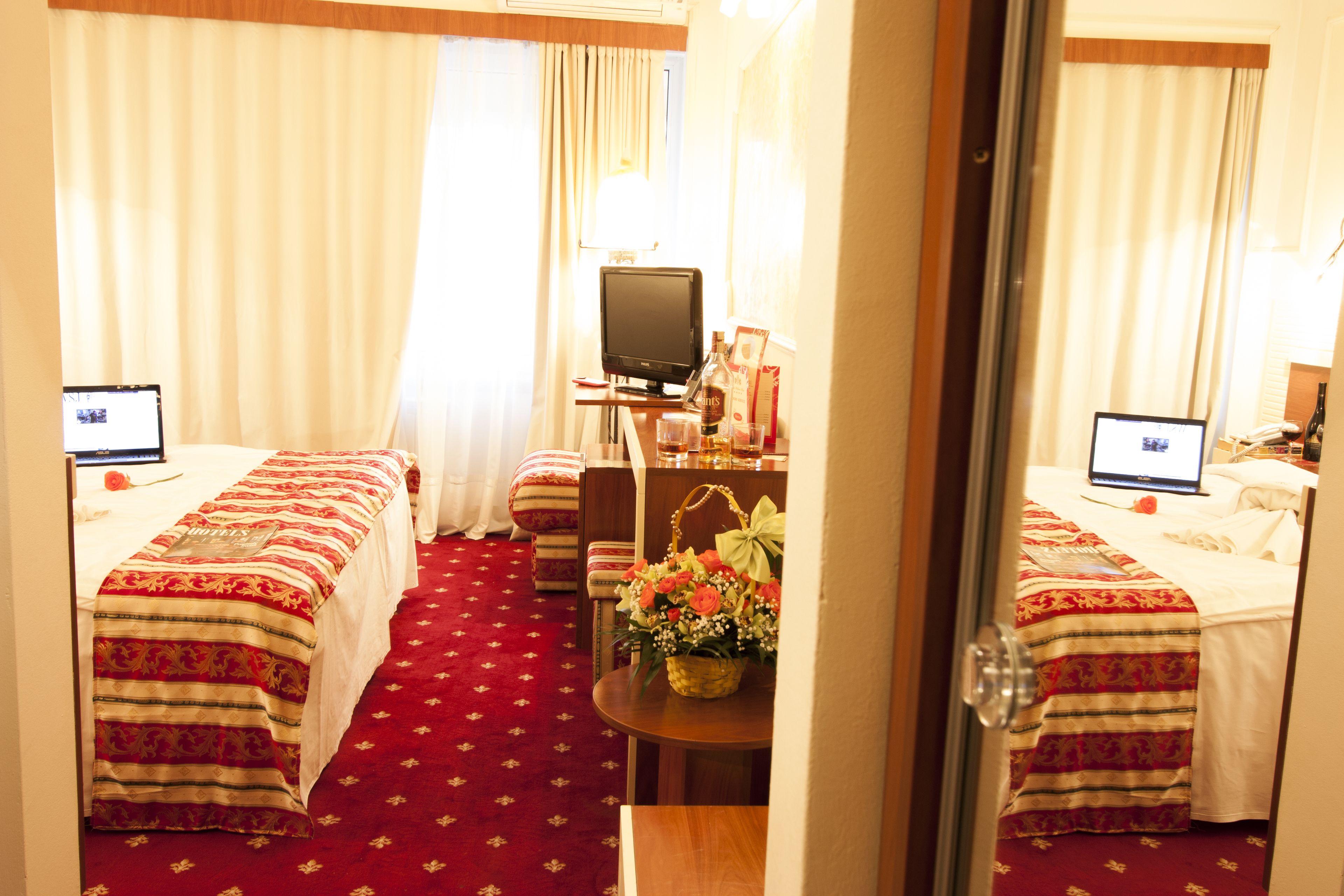 Ein Hotelzimmer mit zwei Betten, einem Schreibtisch mit Computer, einem Fernseher und einem kleinen Tisch mit Blumen.