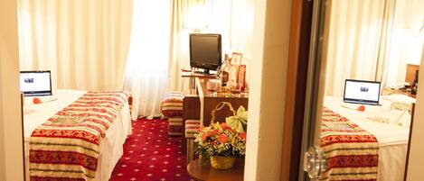Ein Hotelzimmer mit zwei Betten, einem Schreibtisch mit Computer, einem Fernseher und einem kleinen Tisch mit Blumen.