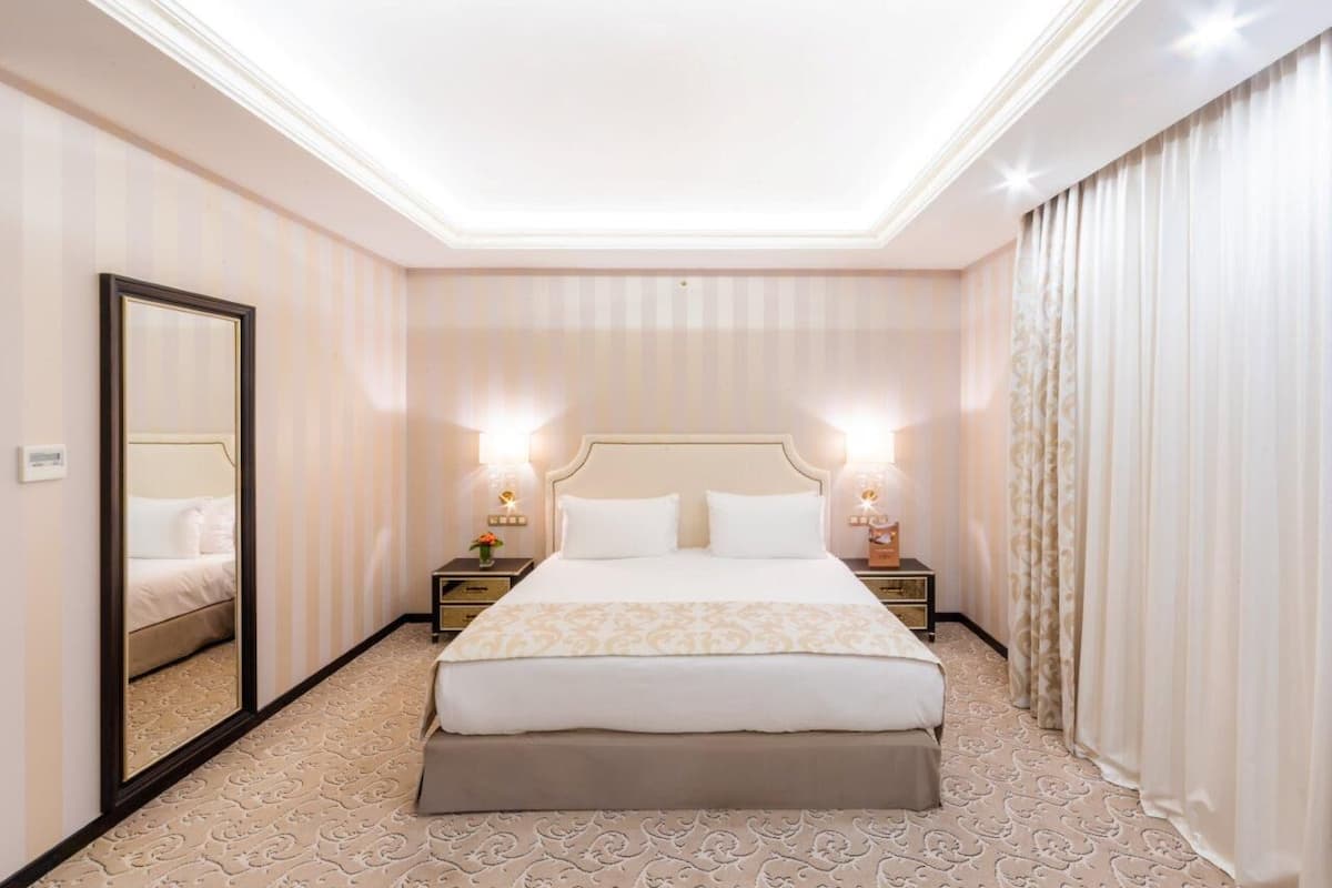Een hotelkamer met een groot bed, twee nachtkastjes met lampen, een spiegel en een kamerbreed tapijt met patroon.