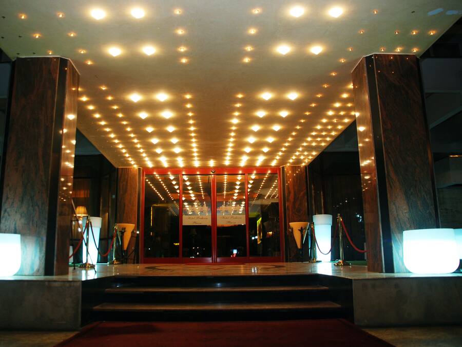 Entrada de hotel con alfombra roja, puerta de cristal y un techo con múltiples luces.
