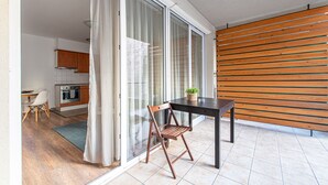 A modern kitchen with wooden cabinets, a dining area with a table and chair, and a sliding glass door leading to a balcony with a wooden slatted wall. - Nova Apartments (Budapest)