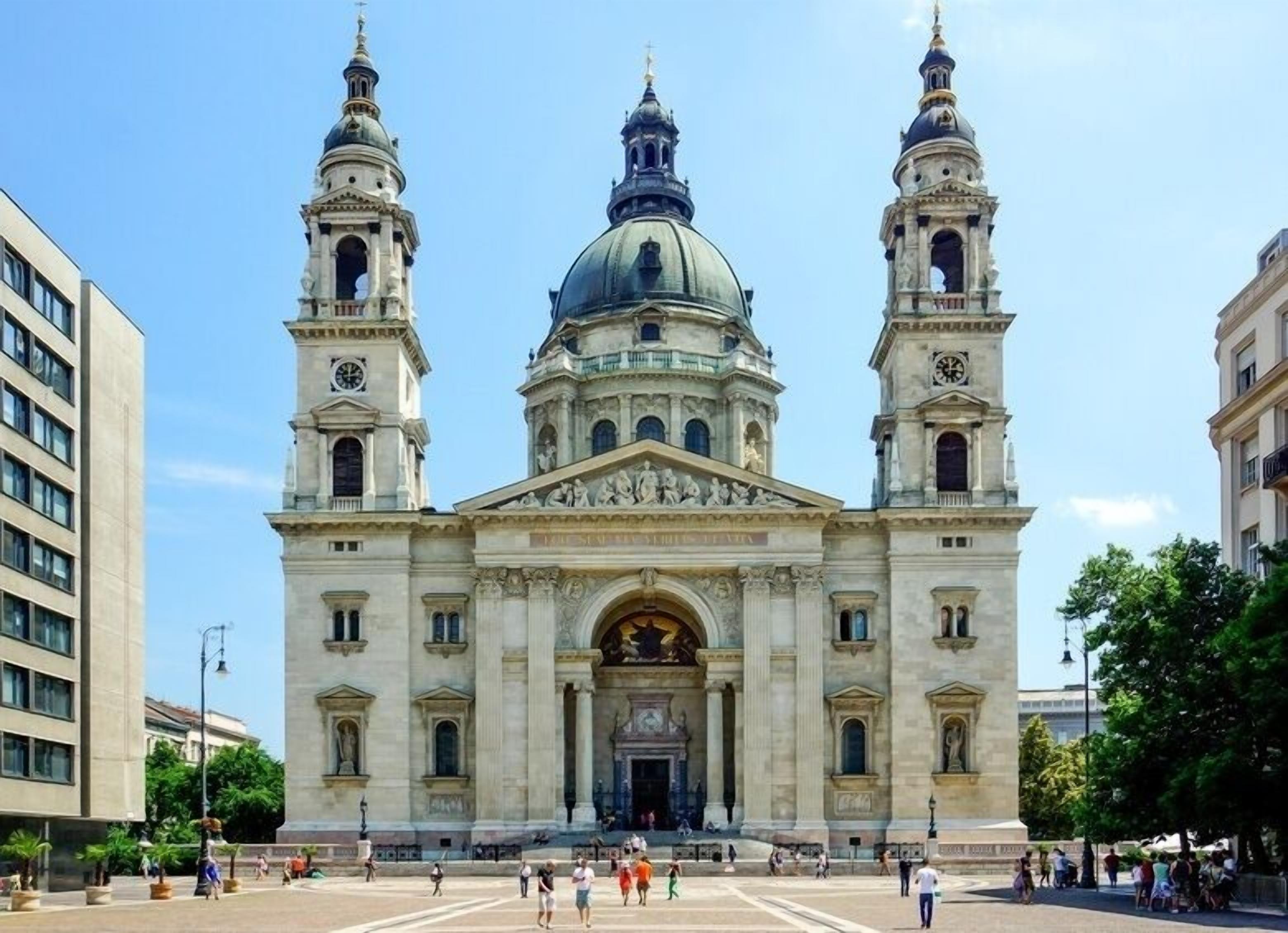 A large, historic building with three towers and a central dome, surrounded by a plaza with people.