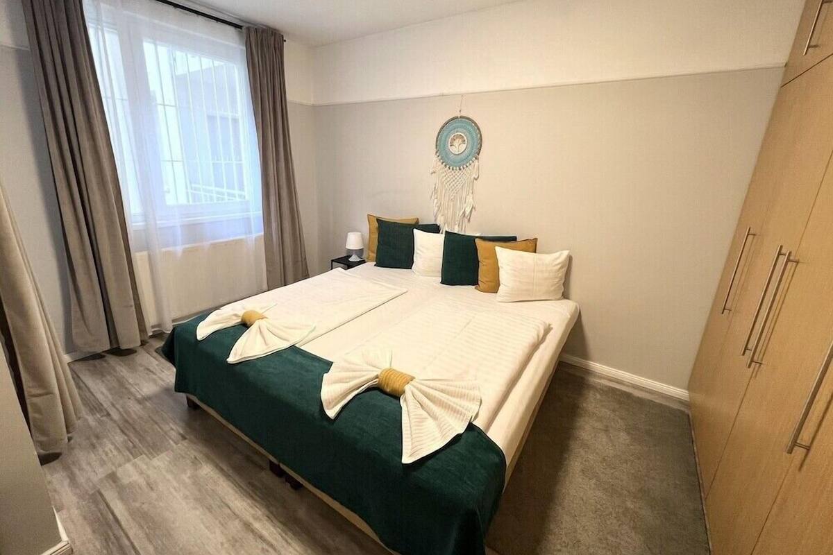 Cama bem arrumada com edredom verde e roupa de cama branca, mesa de cabeceira com abajur, objeto decorativo na parede e guarda-roupa.