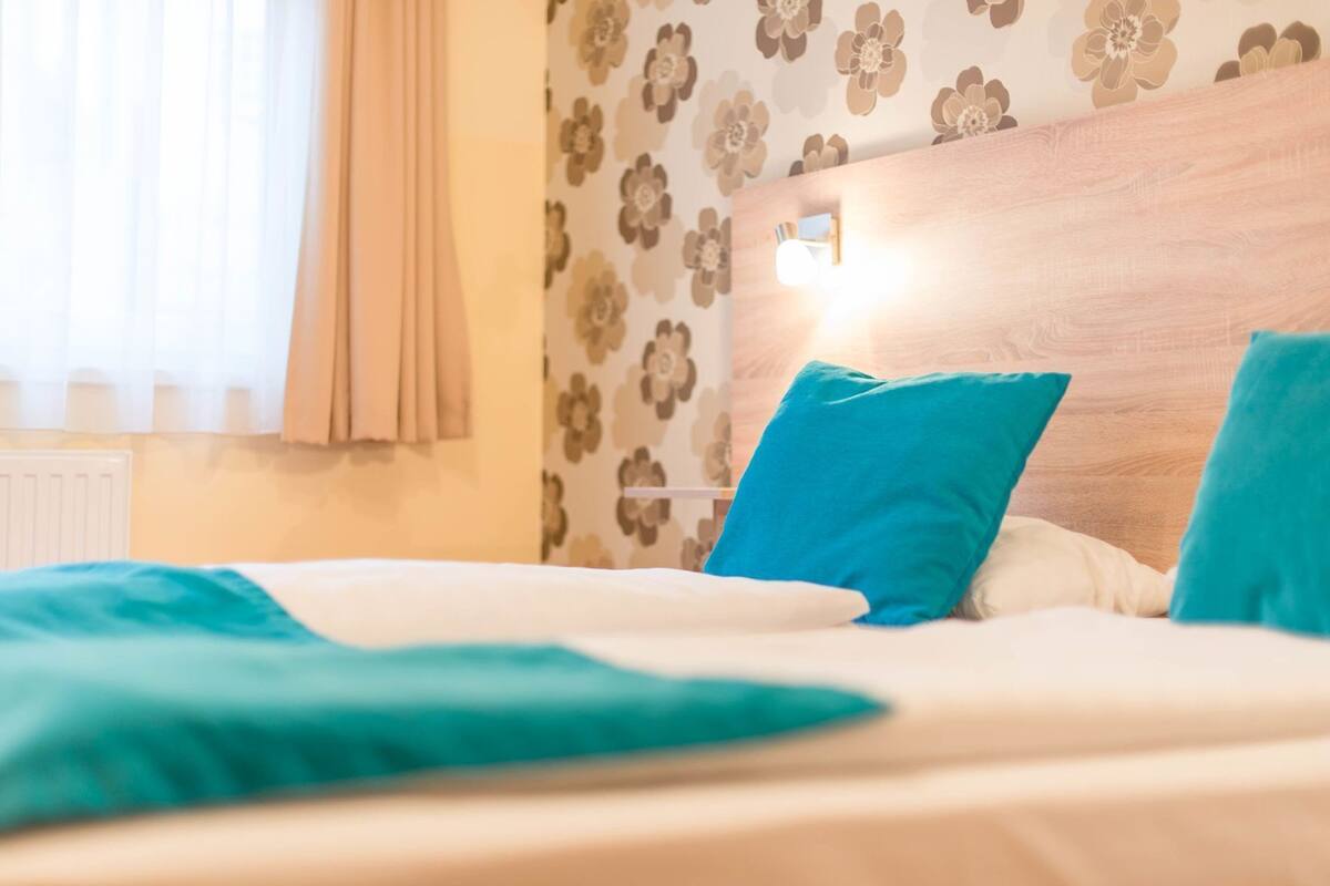 Quarto de hotel com cama, mesa de cabeceira, luminária na parede e papel de parede estampado com flores.