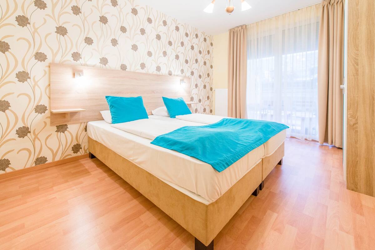 Quarto de hotel com cama, cabeceira, janela com cortinas e piso de madeira.