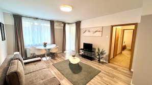 A modern living room with a sofa, a coffee table, a TV, and a dining area with chairs and a table. - Nova Apartments (Budapest)
