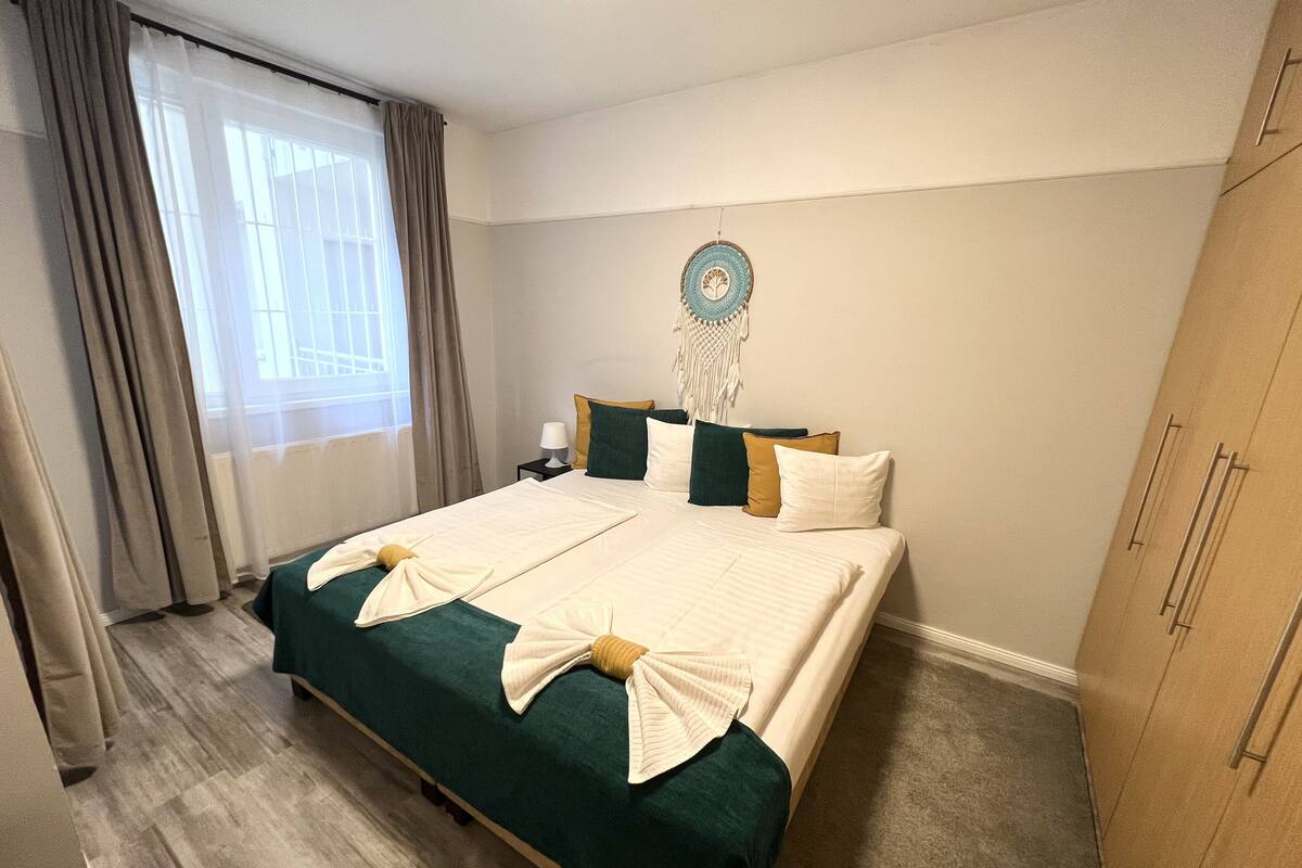 Cama bem arrumada com colcha verde e lençóis brancos, guarda-roupa de madeira e uma decoração de macramé na parede.
