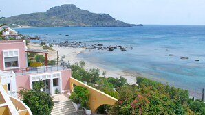 A coastal view with a mountain in the background, a beach with rocks, and a building with a balcony overlooking the sea. - Horizon Beach Hotel (Agios Vasileios)