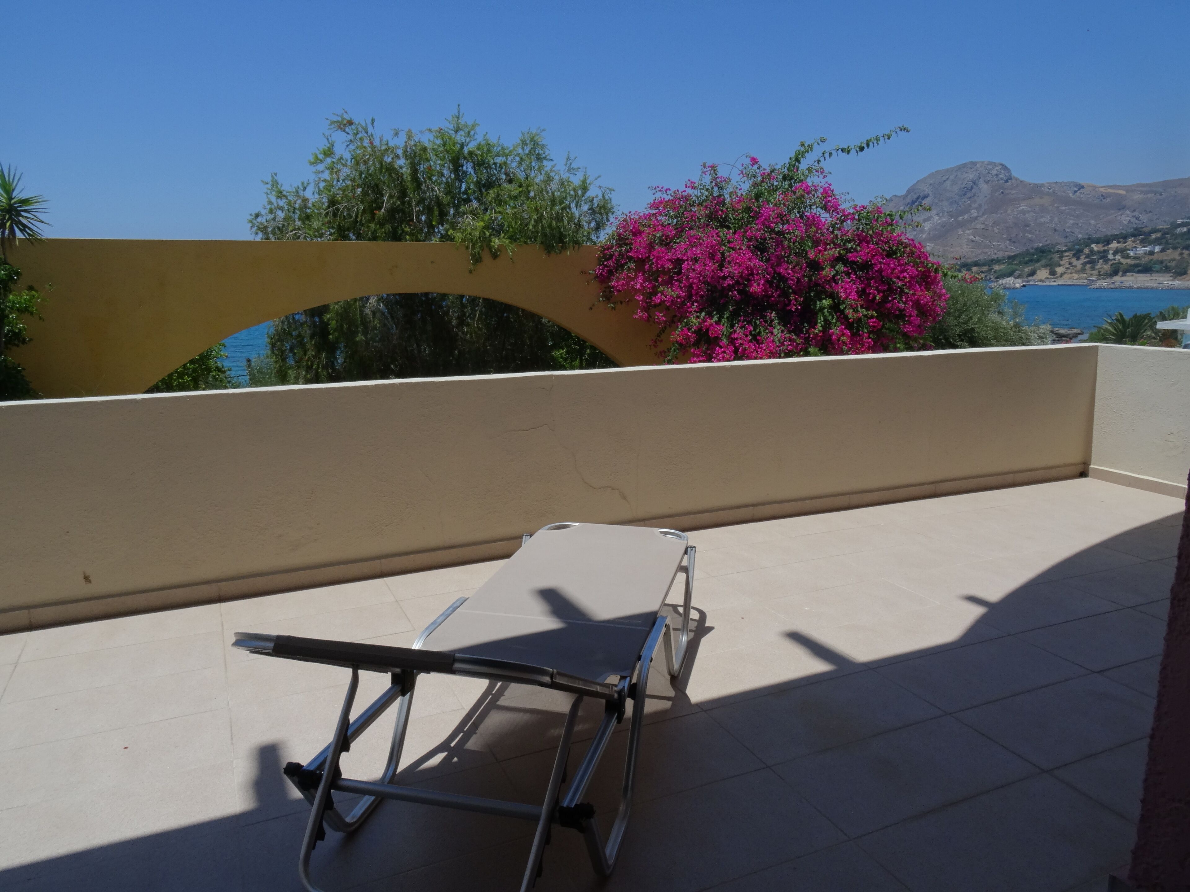 A balcony with a metal chair, a beige wall, and a view of the sea and mountains.