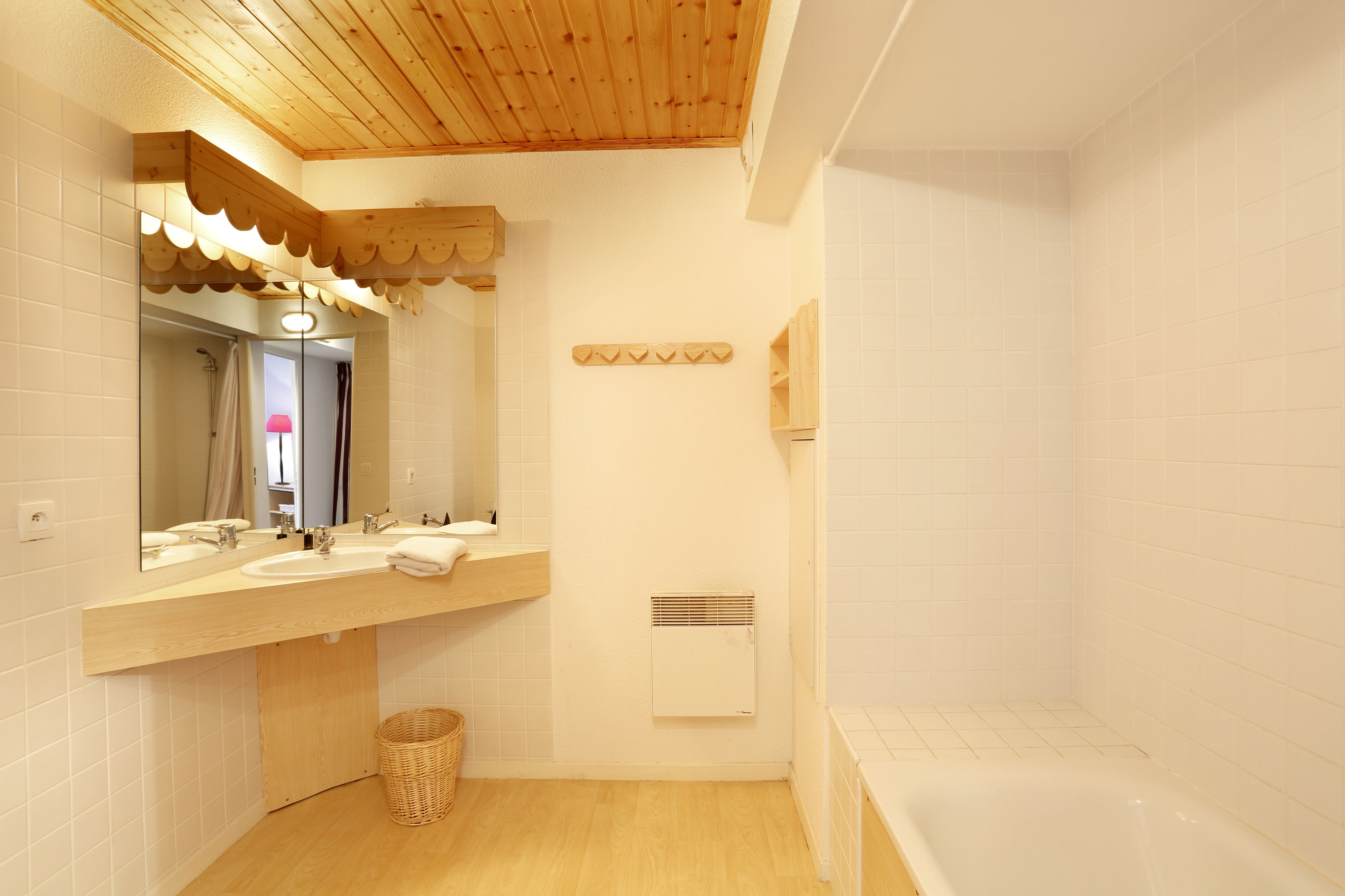 A modern bathroom with wooden ceiling, white tiles, a large mirror, two sinks, a shower, and a white bathtub.