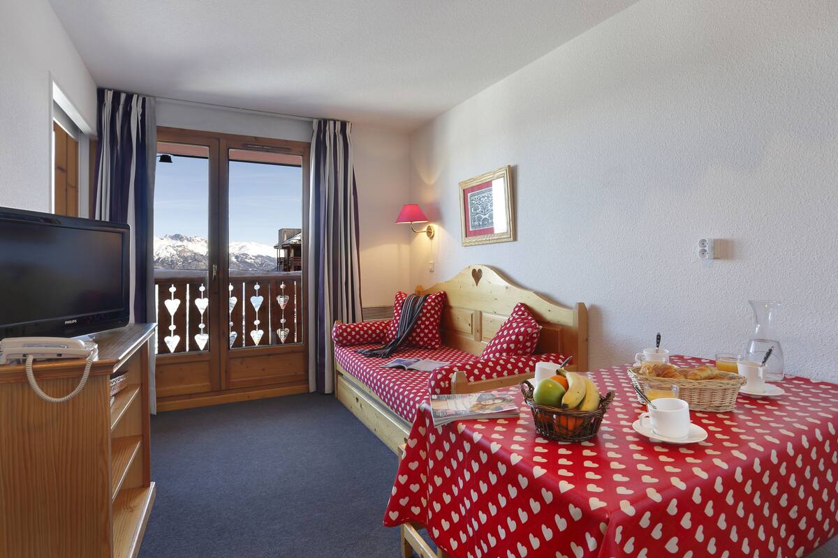 Habitación de hotel con cama, televisión, mesa de desayuno y cesta de fruta, y vistas a las montañas a través de la ventana.