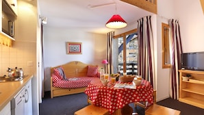 A cozy room with a bed, a dining table with a red tablecloth, a kitchen area with a washing machine, and a television on a wooden stand. - Vacancéole - Résidence Vega (Risoul)