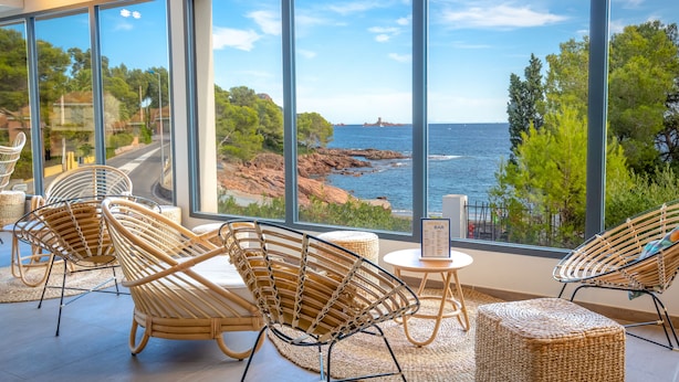 A modern interior with large windows overlooking a coastal landscape, wicker furniture, and a small table with a menu. - SOWELL RÉSIDENCES Les Chênes Verts (Saint-Raphael)
