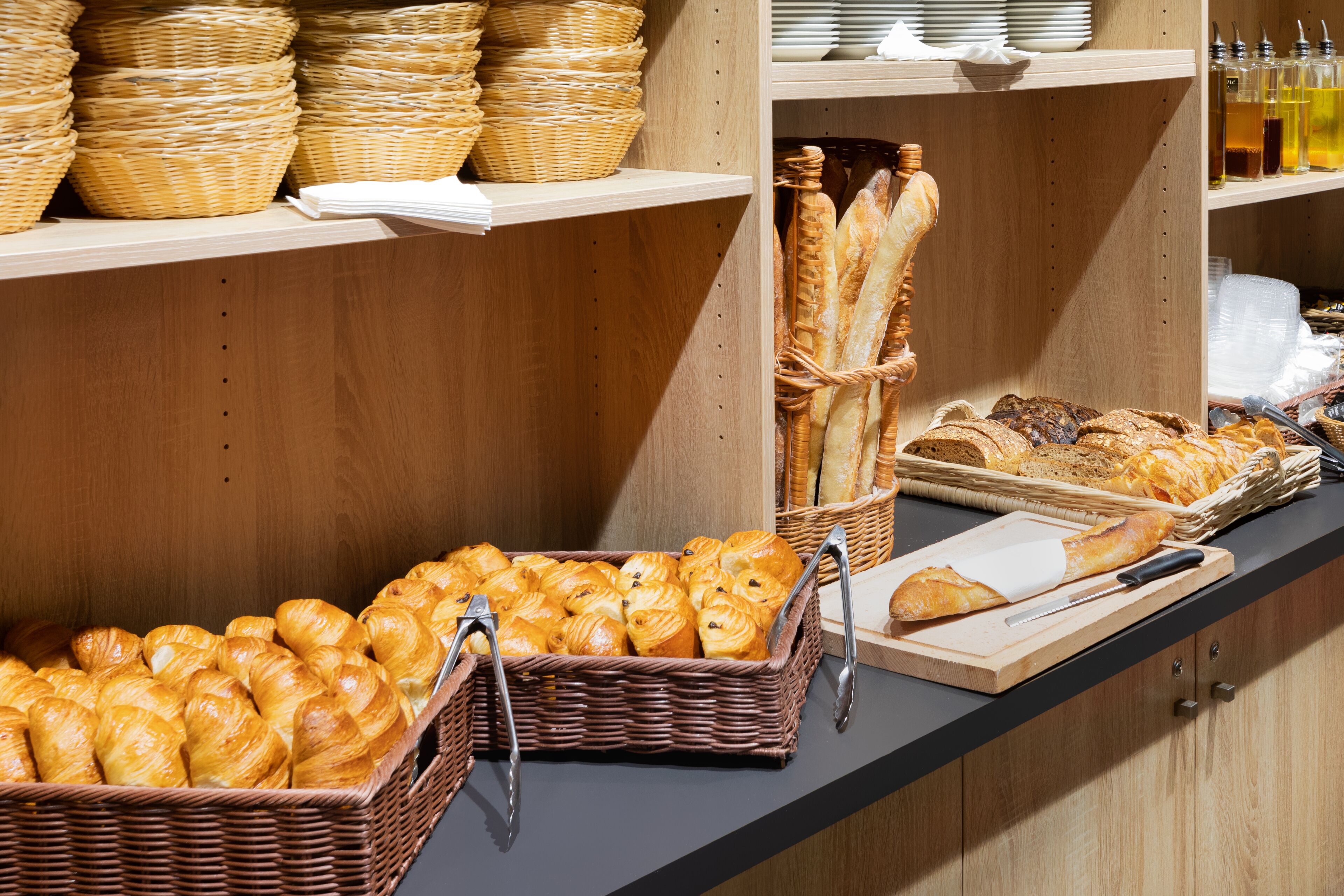 Eine Bäckereiauslage mit Körben voller Croissants und Baguettes, ein Schneidebrett mit Messer sowie Regale mit Tellern und Flaschen.