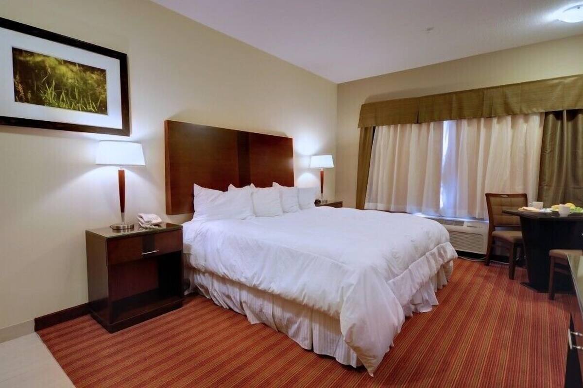 A hotel room with a large bed, two bedside tables with lamps, a small table with a chair, a window with curtains, and a framed picture on the wall.