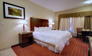 Habitación de hotel con una cama grande, dos mesitas de noche con lámparas, una pequeña mesa con una silla, una ventana con cortinas y un cuadro en la pared.