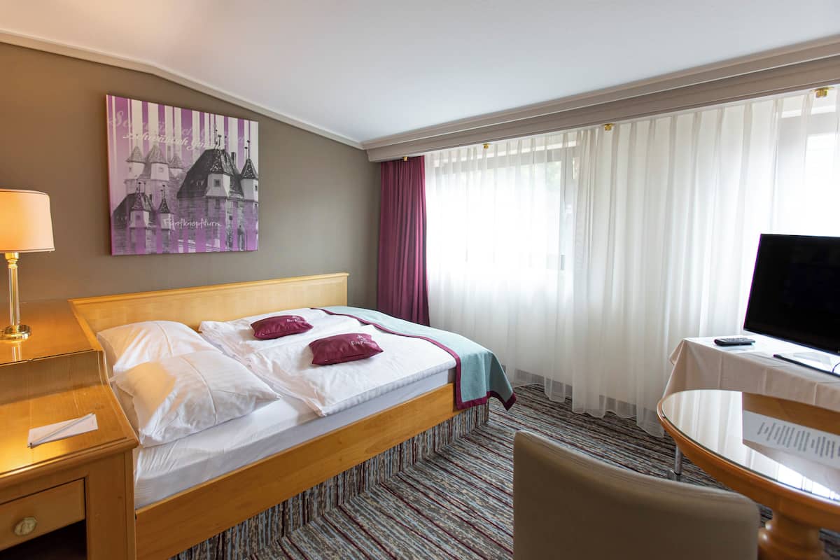 Ein Hotelzimmer mit Bett, Schreibtisch, Stuhl, Fernseher und einem Gemälde an der Wand.