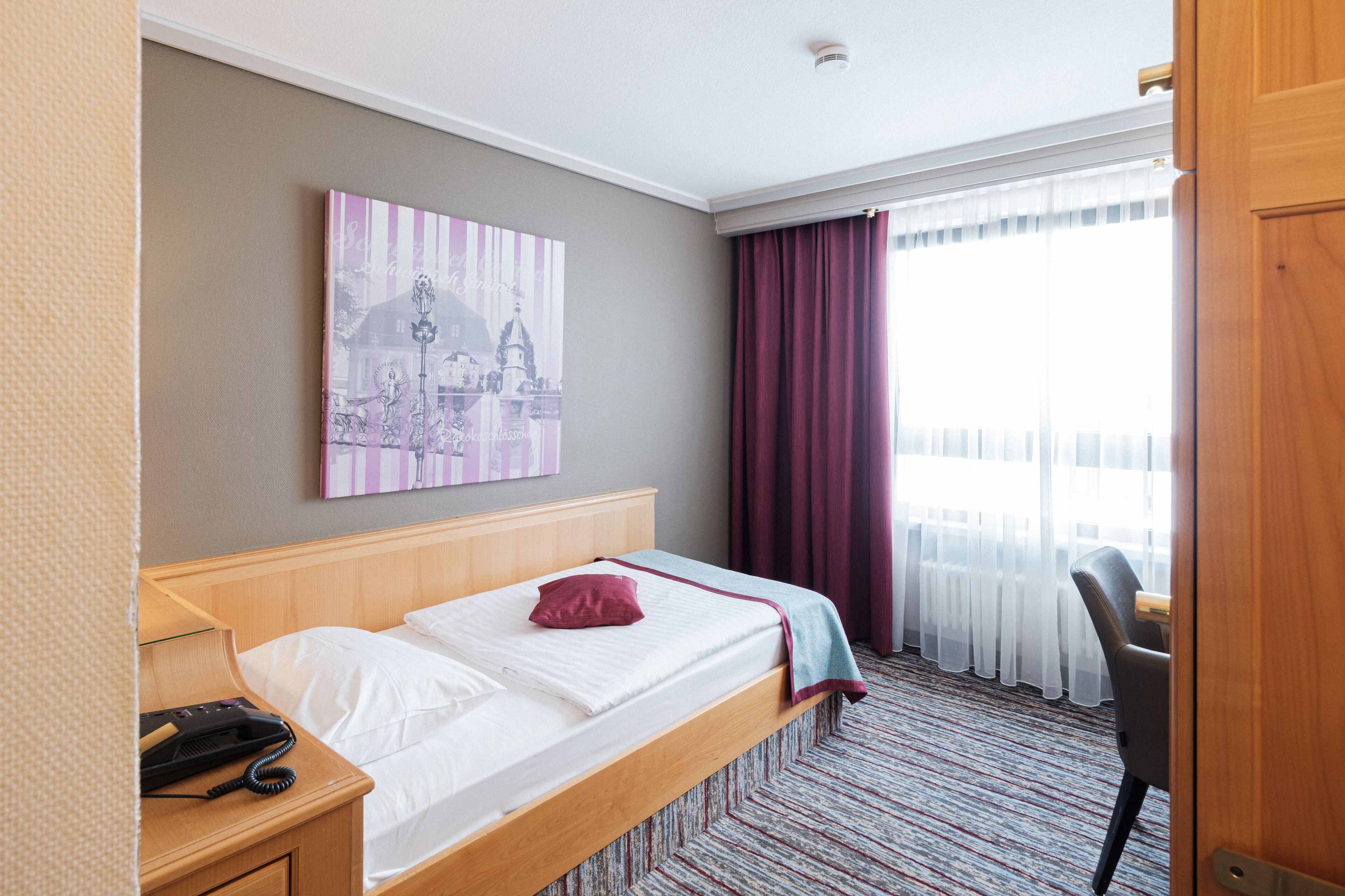 Ein Hotelzimmer mit einem hölzernen Bett, einem Schreibtisch mit Telefon, einem Stuhl, einem Fenster mit transparenten Vorhängen und einem an der Wand hängenden Kunstwerk.